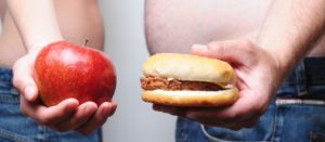 Jóvenes de perfil, uno delgado con una manzana en la mano, y otro obeso con una hamburguesa
